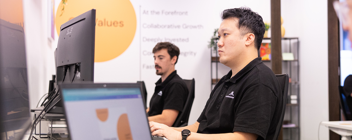 Two AI Advancements developers in black polo shirts working at computer workstations in their office with company values poster visible in the background, representing AI Reporting Agents development team