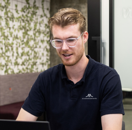 AI Advancements team member with light hair and glasses in a dark blue polo shirt working at a laptop in a modern office with green plant wall, for the About Us page