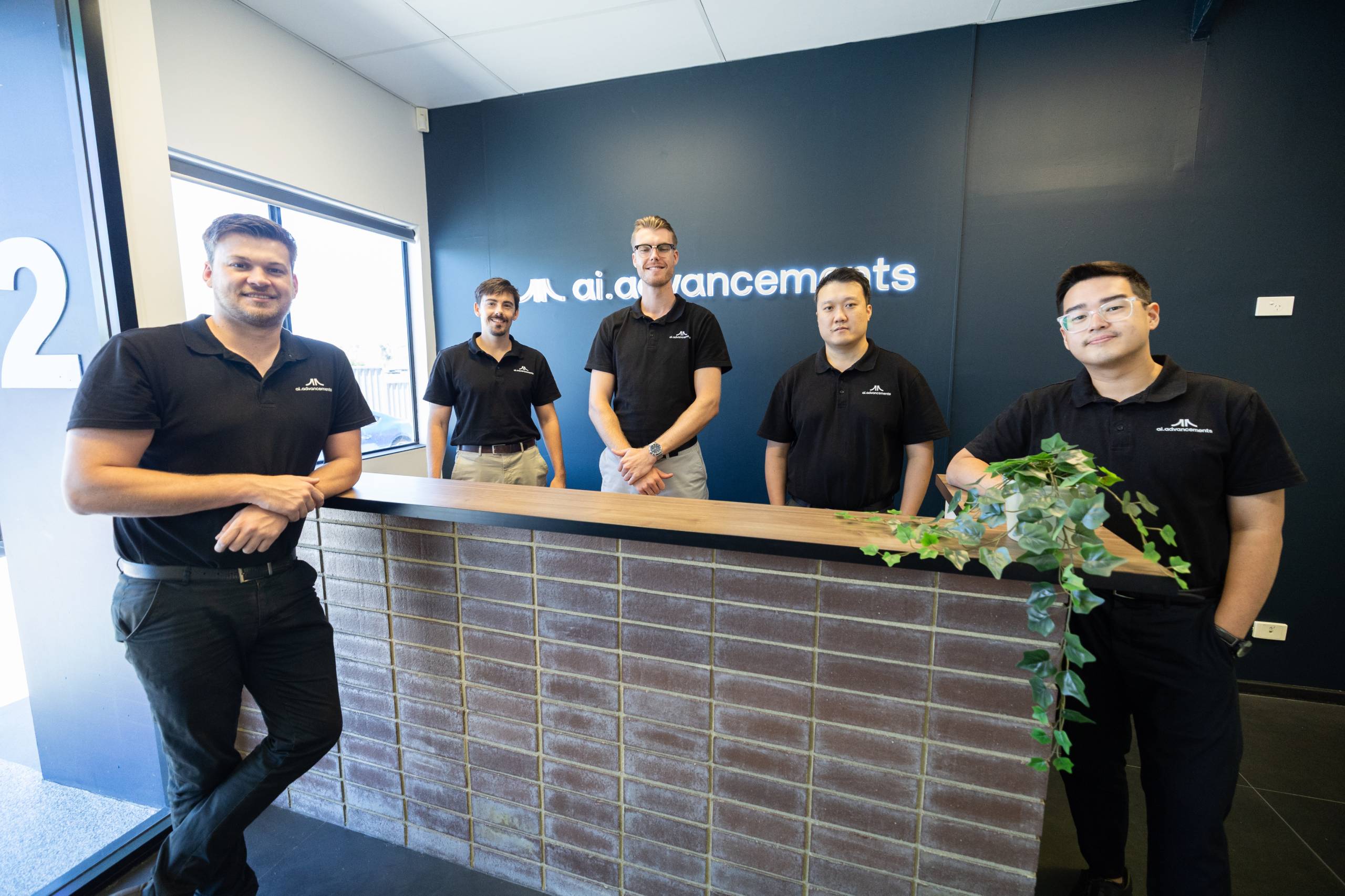 AI Advancements team of five professionals in black polo shirts posing at their office reception desk in front of the dark blue AI Advancements branded company wall