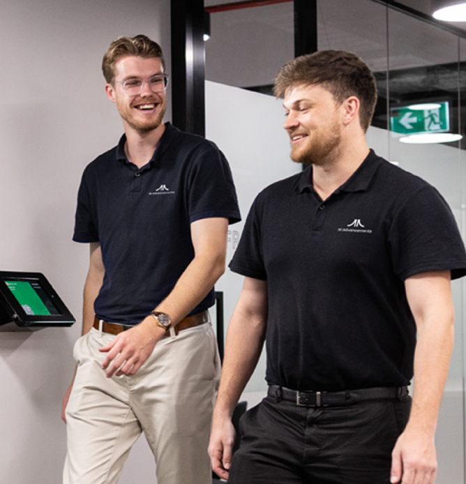 Two young AI Advancements consultants in black polo shirts standing and laughing together in their office, representing the friendly and collaborative team culture on the About Us page
