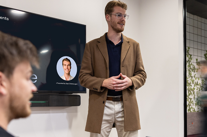 AI Advancements team member in a tan blazer presenting to a group with a large screen showing a profile photo, representing the About Us team introduction and company culture