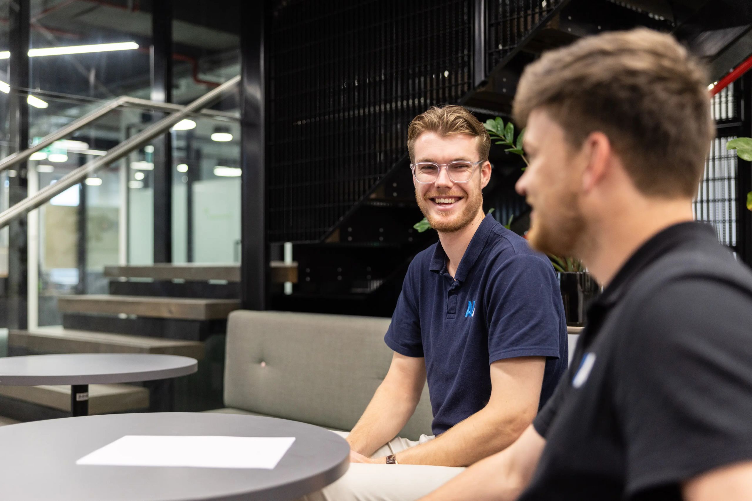 Two professionals engaged in discussion at a modern co-working space, one wearing a navy AI Advancements polo shirt and laughing, photographed in a collaborative AI strategy meeting