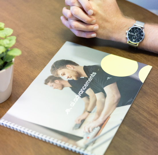 Close-up of hands with watch holding an AI Advancements branded spiral-bound workbook on a wooden table next to a green potted plant, representing AI learning hub course materials