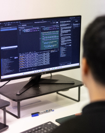 Developer viewed from behind working at a large monitor displaying dark code editor or terminal, representing AI for Developers services