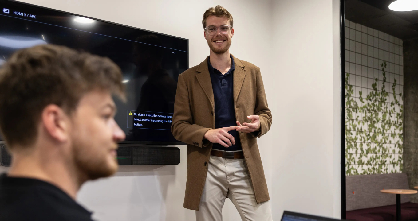 Two AI Advancements team members in a modern office space, one in a tan jacket with glasses smiling and gesturing while presenting to a colleague in the foreground