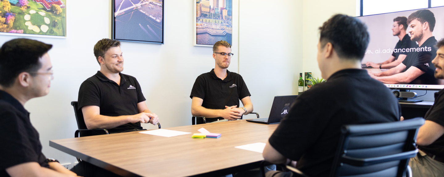 Four AI Advancements team members in black polo shirts having a collaborative meeting around a conference table with an AI Advancements branded screen in the background, representing Phone Agents AI implementation team