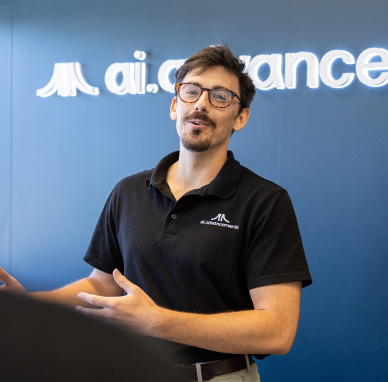 AI Advancements consultant with beard and glasses presenting and gesturing in front of a blue AI Advancements branded backdrop, representing Phone Agents AI services