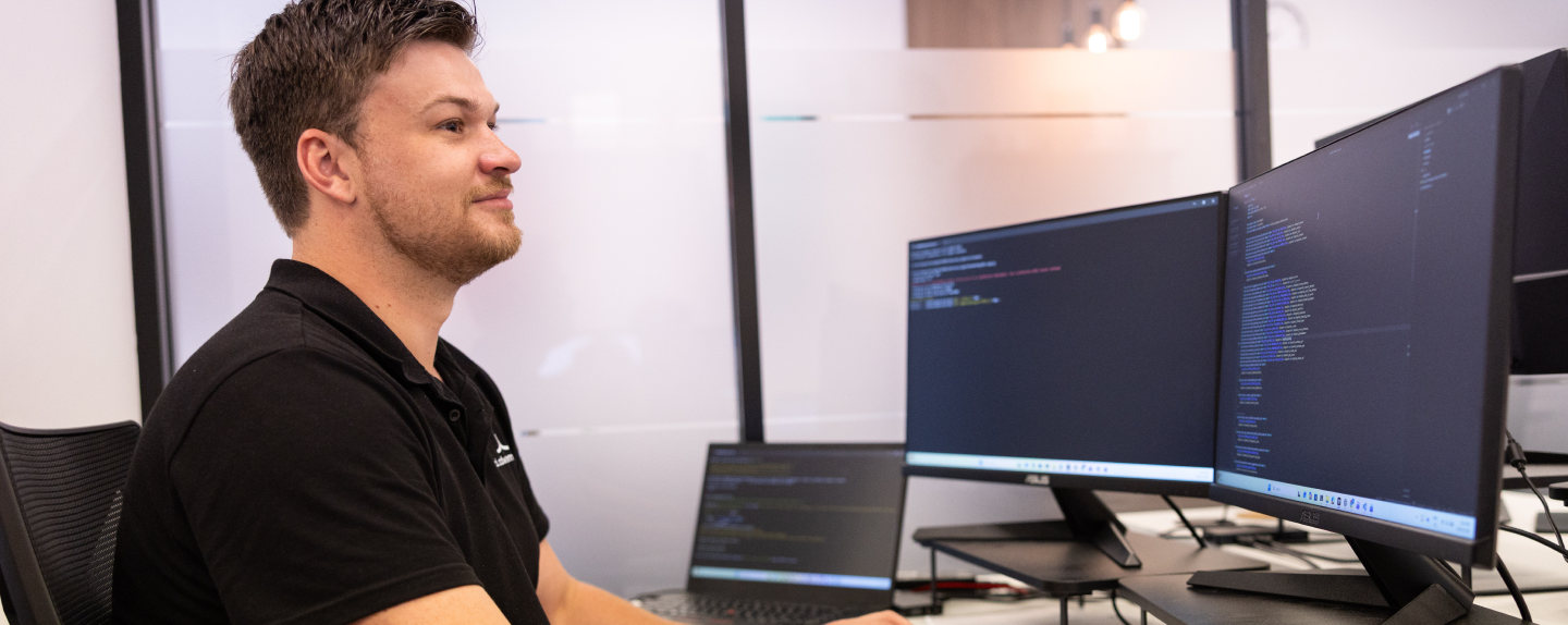 AI Advancements developer in black polo shirt seated at a multi-monitor workstation displaying code, representing AI for Developers programming and development services