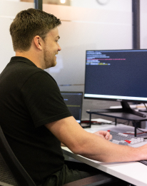 Technical professional in dark shirt working at a multi-monitor computer setup, representing ongoing maintenance and support for AI development