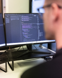Developer working at a large monitor displaying code or terminal, representing the AI automation step to refine, standardise and scale solutions