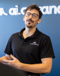 AI trainer with glasses in dark shirt presenting enthusiastically in front of AI Advancements backdrop, representing the step to train your teams and spark ideas