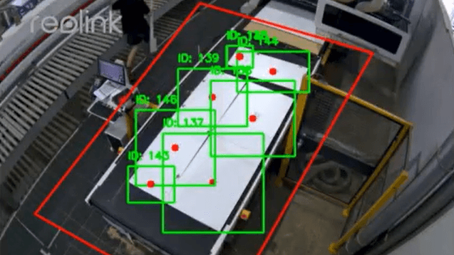 Overhead CCTV camera view from Reolink showing computer vision AI detecting and tracking small manufacturing parts with green ID-numbered bounding boxes and red safety zone boundary on a production workstation