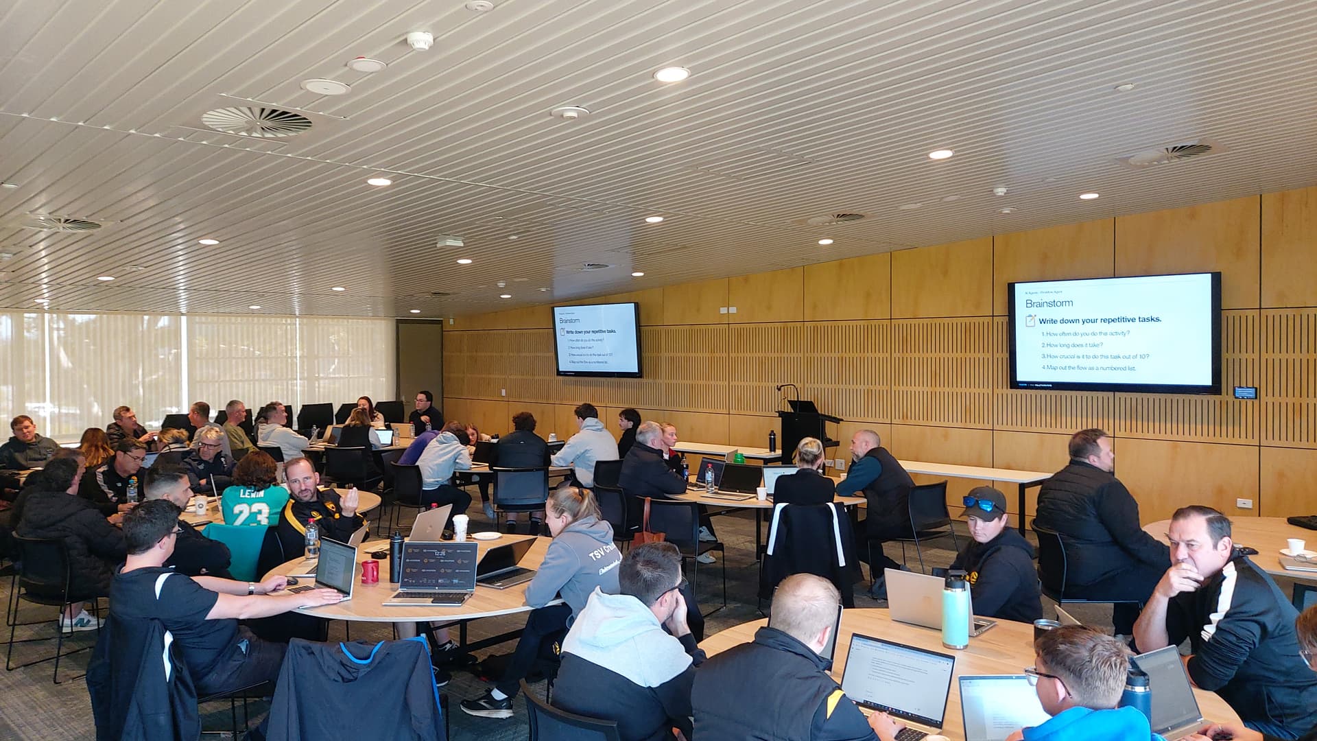 Wide-angle photo of Football West AI training day showing many participants including staff in football jerseys seated at tables with laptops in a modern conference room with presentation screens